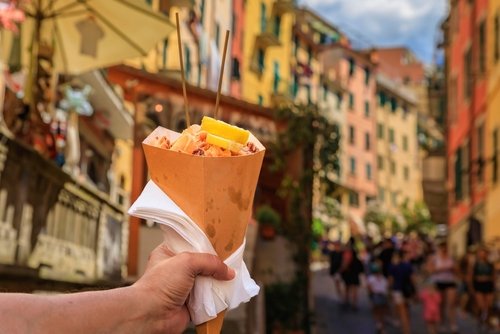 Hand holding a cone of fritto misto, Italian street food mix of fried calamari and shrimp with colorful houses in Riomaggiore, Cinque Terre Italy